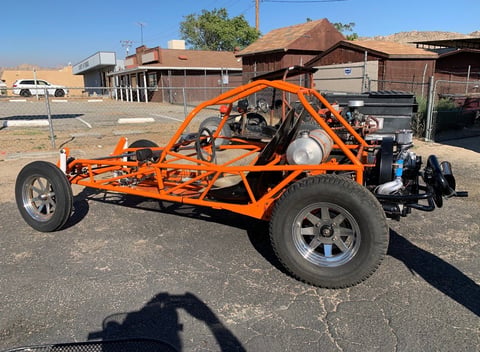 Orange dune buggy with exposed frame parked in a lot with rural buildings in the background