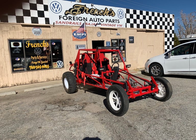Red custom buggy on display outside Frenchs Foreign Auto Parts shop with checkered banner and Volkswagen signage
